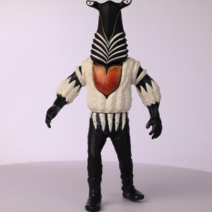 May include: A vintage action figure of a humanoid alien with a black head and body, white chest, and black pants. The figure has a unique design with a tall, zebra-striped head, and a heart-shaped design on the chest.