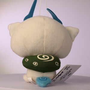 Puede incluir: Un juguete de peluche blanco con dos cuernos azules en la parte superior, un cuello verde con un patr&oacute;n de espirales blancas y una peque&ntilde;a cola azul con un dise&ntilde;o de espiral. Una etiqueta rectangular blanca con texto negro est&aacute; unida al lado.