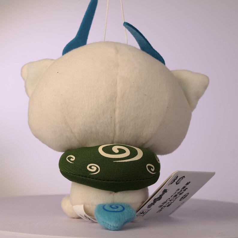 Puede incluir: Un juguete de peluche blanco con dos cuernos azules en la parte superior, un cuello verde con un patr&oacute;n de espirales blancas y una peque&ntilde;a cola azul con un dise&ntilde;o de espiral. Una etiqueta rectangular blanca con texto negro est&aacute; unida al lado.