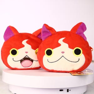 Puede incluir: Dos monederos de peluche con forma de gato. Los monederos son rojos con rasgos faciales blancos y negros, incluyendo grandes ojos con borde amarillo. Las orejas son moradas. Un monedero tiene la boca abierta con lengua rosa. Sobre una superficie blanca.