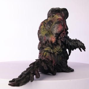 Puede incluir: Escultura detallada de una criatura con una apariencia org&aacute;nica texturizada. La figura es principalmente verde oscuro y negra, con acentos de rojo y amarillo. La criatura est&aacute; sentada con una larga cola.