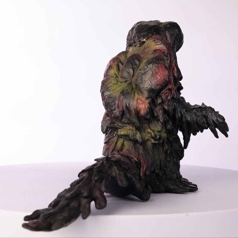 Puede incluir: Escultura detallada de una criatura con una apariencia org&aacute;nica texturizada. La figura es principalmente verde oscuro y negra, con acentos de rojo y amarillo. La criatura est&aacute; sentada con una larga cola.