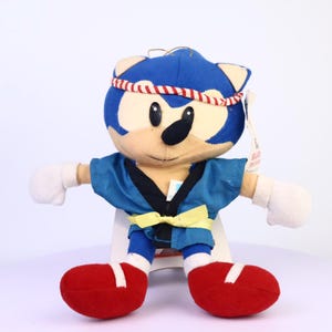 May include: A plush toy of Sonic the Hedgehog in a blue karate uniform with a black belt and a yellow bow. The toy has white gloves, red shoes, and a red and white headband. The toy is standing with its arms outstretched.