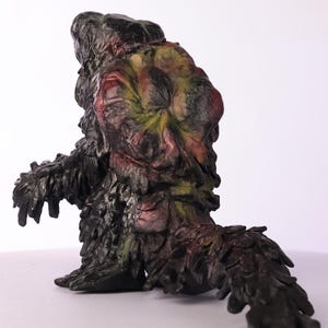 Puede incluir: Una figura esculpida de una criatura con un cuerpo oscuro texturizado y una cabeza con tonos rojos, amarillos y verdes. La figura tiene una cola y parece ser un monstruo o un personaje de fantas&iacute;a. La escultura est&aacute; sobre un fondo blanco.