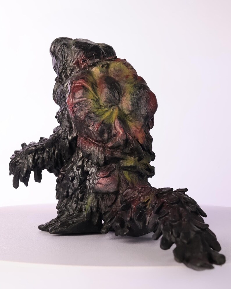 Puede incluir: Una figura esculpida de una criatura con un cuerpo oscuro texturizado y una cabeza con tonos rojos, amarillos y verdes. La figura tiene una cola y parece ser un monstruo o un personaje de fantas&iacute;a. La escultura est&aacute; sobre un fondo blanco.