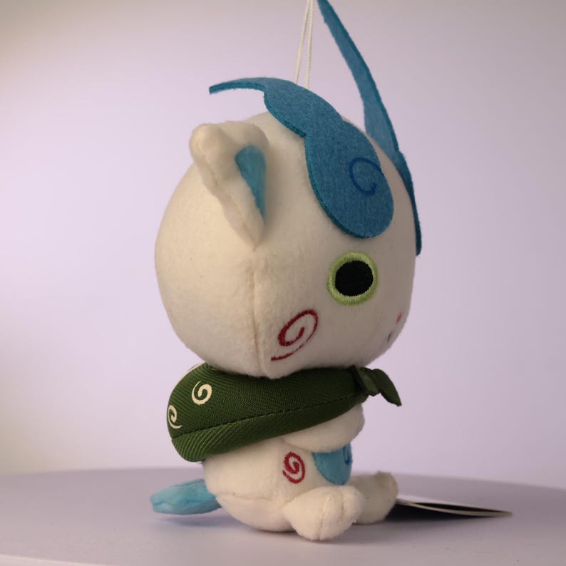 Puede incluir: Un juguete de peluche blanco con una cabeza grande, orejas de fieltro azul en forma de llama, un ojo negro con borde verde y marcas de espirales rojas en las mejillas. La figura lleva una capa de tela verde oscuro con patrones de espirales blancos.