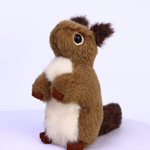 May include: A plush squirrel toy in shades of brown and cream. The stuffed animal has a fluffy tail, small ears, and black eyes. The squirrel is standing upright, with its paws together, and is a cute, soft toy.