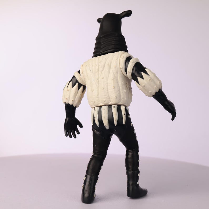 May include: A black and white figurine of a character with a tall, hooded head and a white jacket with black accents. The figure is wearing black gloves, pants, and boots. The jacket has a quilted texture and the figure is posed from the back.
