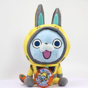 May include: A yellow and blue plush toy with bunny ears wearing a yellow and black spacesuit. The toy is holding a red and blue Yo-kai Medal with a picture of a yellow and black Yo-kai on it.