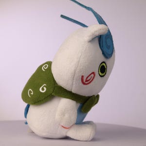 Puede incluir: Un peluche blanco que presenta una pieza trasera similar a un caparaz&oacute;n verde con bordados de espirales blancas, una antena azul curvada, un ojo verde visible con pupila negra y un detalle de espiral roja en la mejilla del juguete.