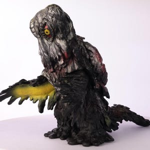 Puede incluir: Figura detallada de una criatura con un cuerpo texturizado gris oscuro y negro. La criatura tiene un ap&eacute;ndice amarillo y verde, un &aacute;rea roja y un solo ojo amarillo. La figura est&aacute; sobre una superficie blanca.