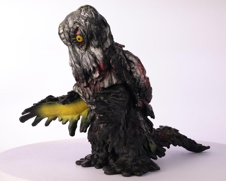 Puede incluir: Figura detallada de una criatura con un cuerpo texturizado gris oscuro y negro. La criatura tiene un ap&eacute;ndice amarillo y verde, un &aacute;rea roja y un solo ojo amarillo. La figura est&aacute; sobre una superficie blanca.