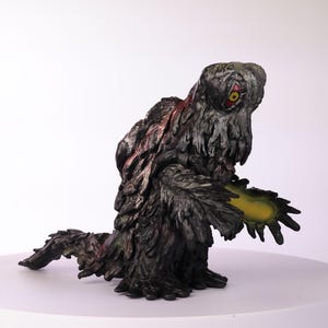 Puede incluir: Figura detallada de una criatura con un cuerpo oscuro y texturizado y un solo ojo grande con iris rojo. La figura tiene un brillo amarillo que emana de su mano. La paleta de colores general es oscura con acentos rojos y amarillos.