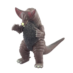 May include: A brown monster figurine with a textured body, large horns with red markings, and a long tail. The toy has a black chest and is posed with its arms raised, ready for action. The figurine is a collectible toy.
