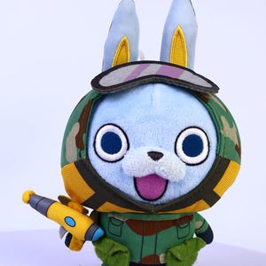 May include: A soft toy rabbit dressed in a camouflage uniform, a visor, and holding a yellow and grey toy weapon. The rabbit has blue fur, large blue eyes, and a pink tongue. The toy is designed with a cartoonish aesthetic.