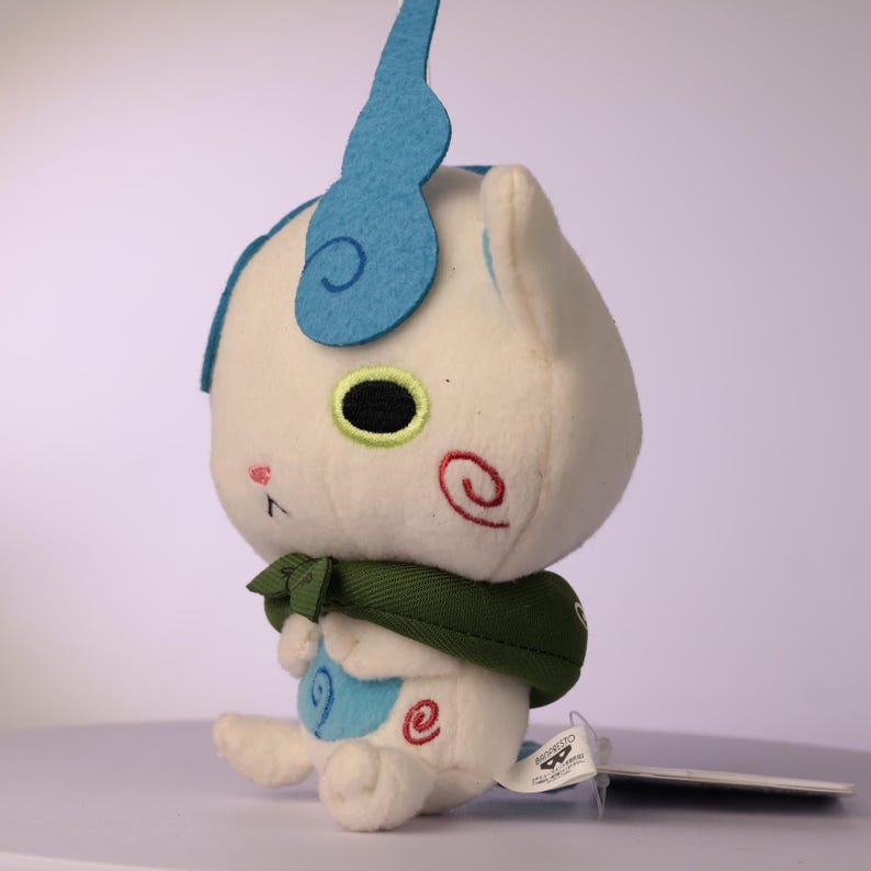 Puede incluir: Un juguete de peluche blanco con una gran cresta de fieltro azul en forma de llama sobre la cabeza, un cuello de tela verde y detalles en espiral bordados en rojo y azul en las mejillas y el vientre, con grandes ojos negros y borde verde.
