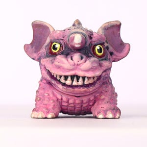 May include: A pink and purple monster figurine with large ears, three eyes, and a wide grin revealing sharp teeth. The creature has a textured, bumpy skin and is posed in a seated position. The eyes are yellow with red irises.
