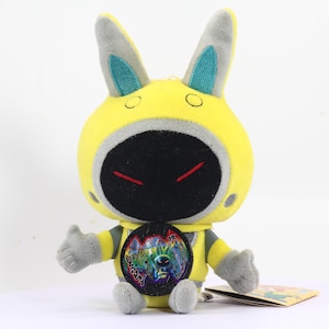 May include: Yellow plush toy with bunny ears, a black face with red eyes, and a round yellow badge with a black and white image of a creature. The toy is sitting on a white surface.