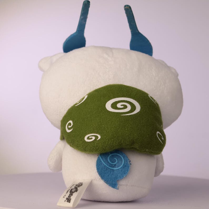 Puede incluir: Un peluche blanco con dos antenas azules erguidas, una pieza trasera verde similar a un caparaz&oacute;n decorada con patrones de espirales blancas y una peque&ntilde;a cola azul con patr&oacute;n de espiral. Una etiqueta de tela blanca con texto negro est&aacute; unida al borde inferior.