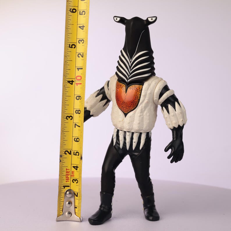 May include: A vintage action figure of a creature with a black head and body, white shirt, and orange heart-shaped chest. The figure is approximately 5 inches tall. The figure has a zebra-like pattern on its head and chest.