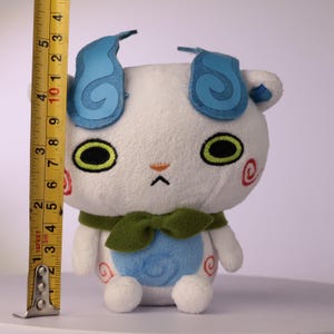 Puede incluir: Un peluche blanco con orejas en forma de llama azul, ojos verdes, una bufanda verde y una mancha azul en el vientre. Tiene espirales rojas en las mejillas y caderas. Una cinta m&eacute;trica amarilla muestra una altura de unos 11,5 cent&iacute;metros.