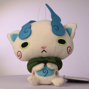 Puede incluir: Un juguete de peluche blanco con una cabeza grande, dos orejas de fieltro azul en forma de llama, ojos ovalados negros con bordes verde lima y una peque&ntilde;a nariz rosa. Lleva una capa de tela verde oscuro y bordados de espirales rojas en las mejillas.