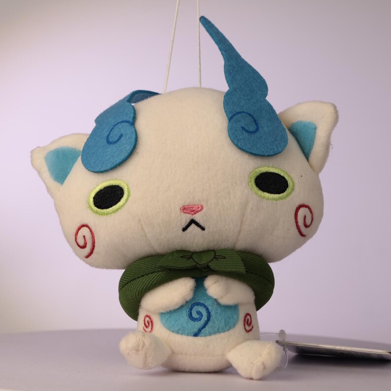 Puede incluir: Un juguete de peluche blanco con una cabeza grande, dos orejas de fieltro azul en forma de llama, ojos ovalados negros con bordes verde lima y una peque&ntilde;a nariz rosa. Lleva una capa de tela verde oscuro y bordados de espirales rojas en las mejillas.