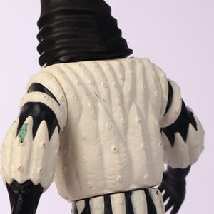 May include: A vintage figurine with a black and white color scheme. The figure has a black head covering and black gloves. The white torso has a textured pattern with small, raised dots. The sleeves are white with black accents. The lower portion of the figure has black and white stripes.