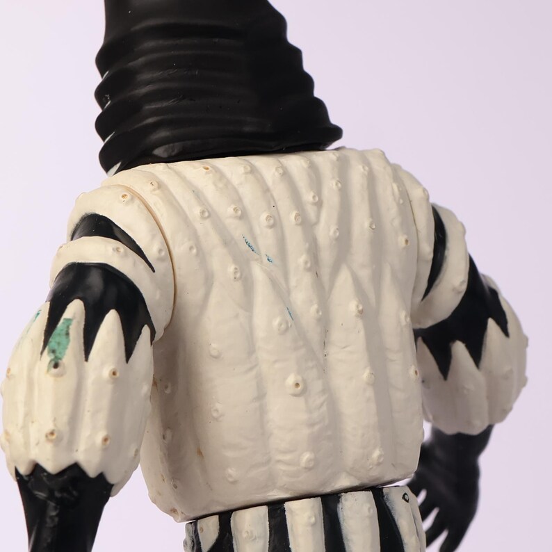 May include: A vintage figurine with a black and white color scheme. The figure has a black head covering and black gloves. The white torso has a textured pattern with small, raised dots. The sleeves are white with black accents. The lower portion of the figure has black and white stripes.