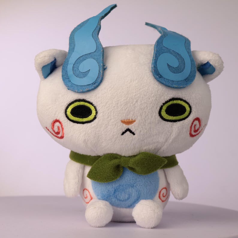 Puede incluir: Un peluche blanco con dos orejas en forma de llama azul, grandes ojos verdes con pupilas negras y una peque&ntilde;a nariz naranja. Presenta espirales rojas en las mejillas y la parte inferior del cuerpo, un parche circular azul en el vientre y una bufanda verde.