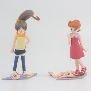May include: Two anime figurines, one with brown hair and a yellow and black outfit, the other with red hair and a red dress. Both figurines are standing on a yellow and black game board.