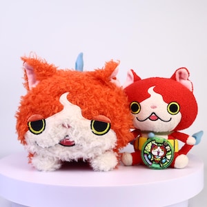 May include: Two plush cat toys. One is orange with white accents and large black eyes. The other is red with white accents and yellow eyes. A colorful medallion is next to the red cat toy.
