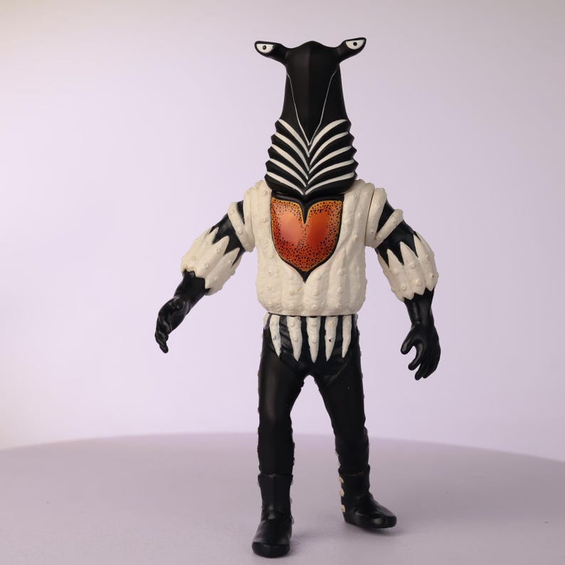 May include: A vintage action figure of a humanoid alien with a black head and body, white chest, and black pants. The figure has a unique design with a tall, zebra-striped head, and a heart-shaped design on the chest.