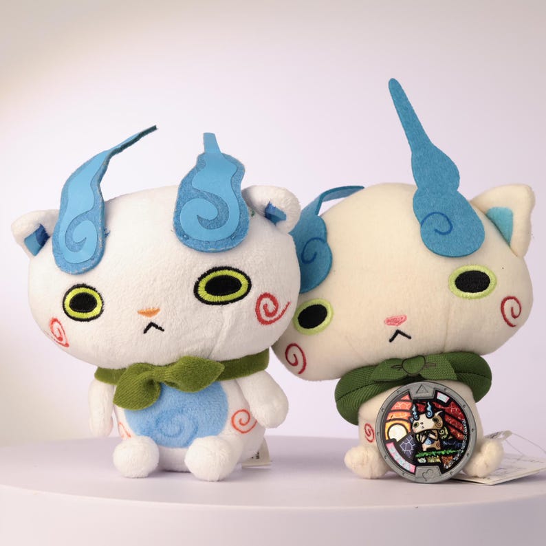 Puede incluir: Dos peluches blancos de cabeza grande, orejas con forma de llama azul y bufandas verdes. Cada figura tiene ojos amarillos grandes, espirales rojas en las mejillas y un parche circular azul en el vientre. Uno sostiene una medalla circular con gr&aacute;fico.