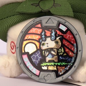 Puede incluir: Una medalla circular de Yo-kai Watch con un marco de color plateado y formas geom&eacute;tricas en relieve. El centro muestra una colorida ilustraci&oacute;n estilo vidriera de una peque&ntilde;a criatura beige con llamas azules sobre un fondo vibrante.