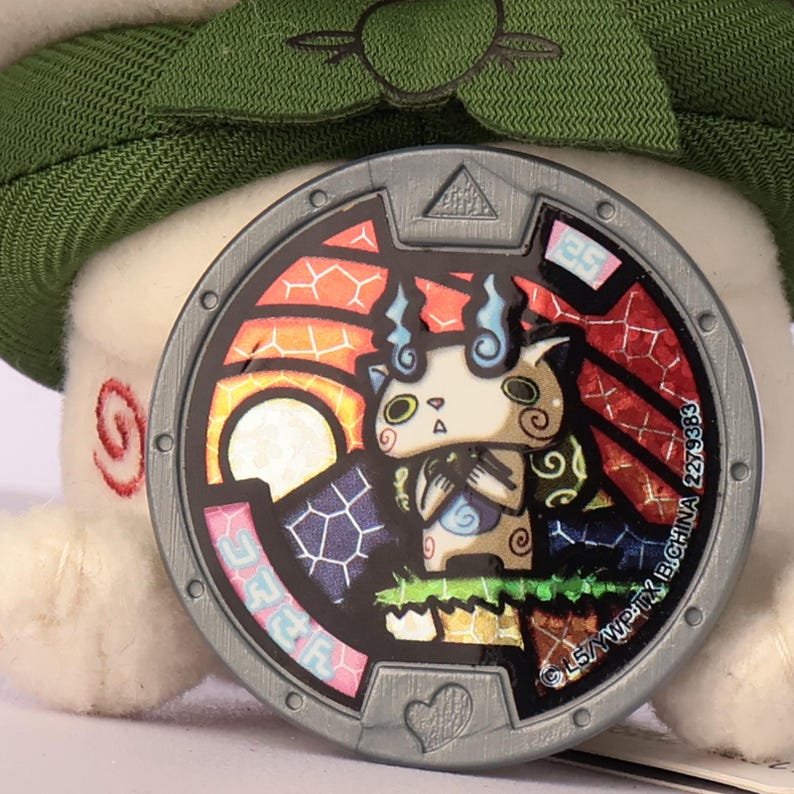 Puede incluir: Una medalla circular de Yo-kai Watch con un marco de color plateado y formas geom&eacute;tricas en relieve. El centro muestra una colorida ilustraci&oacute;n estilo vidriera de una peque&ntilde;a criatura beige con llamas azules sobre un fondo vibrante.