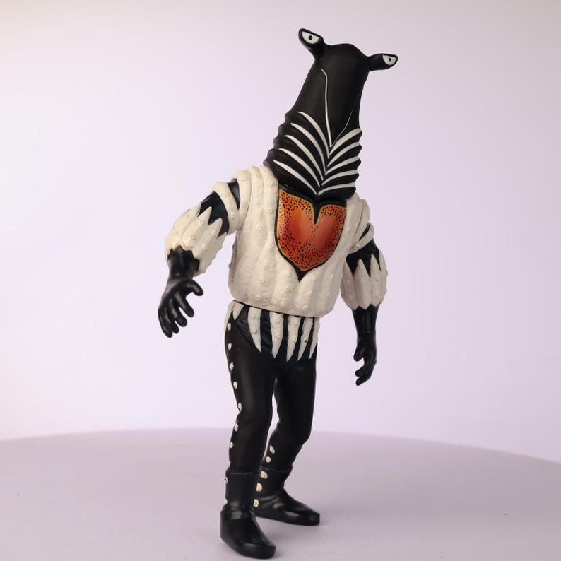 May include: A black and white figurine of a creature with a zebra-like head and a white and black outfit. The figure has a white top with black accents and a red heart-shaped design. The pants are black with white details.