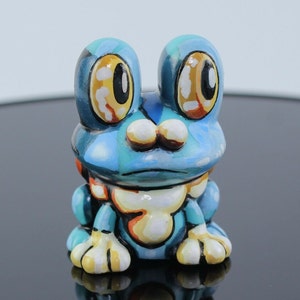 May include: A hand-painted ceramic frog figurine in shades of blue, teal, and white. The frog has large, expressive eyes with yellow and black details. Its belly is cream-colored with orange accents, and its feet are white with yellow stripes.