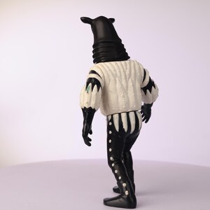 May include: A black and white figurine of a creature with a tall, cylindrical head and a white textured shirt. The figure has black gloves, pants, and boots. The shirt has black accents on the sleeves and a striped pattern on the back.