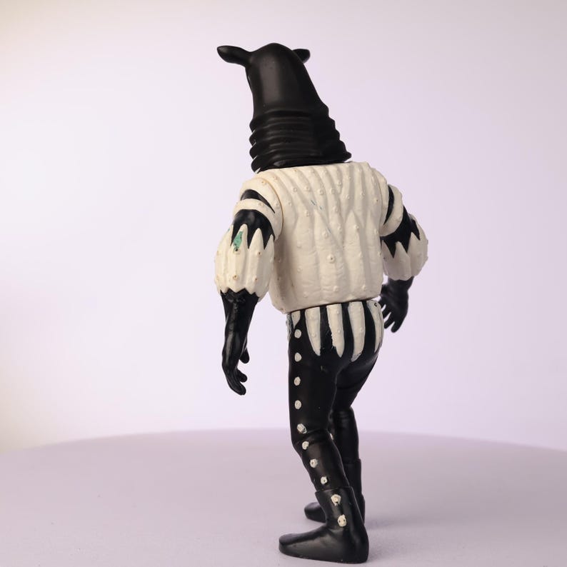 May include: A black and white figurine of a creature with a tall, cylindrical head and a white textured shirt. The figure has black gloves, pants, and boots. The shirt has black accents on the sleeves and a striped pattern on the back.