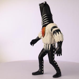 May include: A black and white figurine of a creature with a long neck and a white and orange chest. The figure is wearing a white jacket with black accents and black pants with white dots. The boots are black.