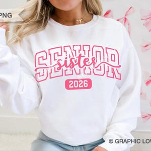 May include: White sweatshirt with the words "SENIOR sister 2026" in pink. The word "SENIOR" is in a block letter font, and "sister" is in a cursive font. The number "2026" is in a pink rectangle.
