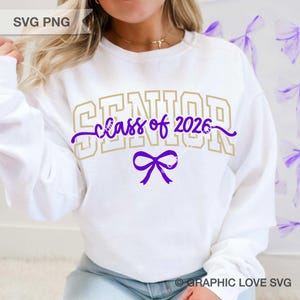 Puede incluir: Sudadera blanca con el texto "SENIOR class of 2026" en dorado y morado. Un lazo morado est&aacute; debajo del texto. La sudadera es de cuello redondo y manga larga.