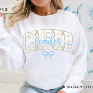 Peut inclure: Sweat-shirt blanc avec le texte "CHEERleader" en lettres dorées et bleues avec un nœud bleu en dessous. Le texte est dans une police de caractères de style universitaire.