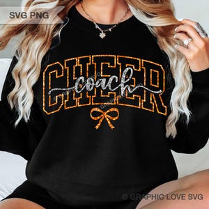 May include: A black long-sleeve sweatshirt featuring the word CHEER in large, orange glitter block letters. The word coach is written in silver glitter script across the center, and a small orange glitter bow is positioned below the text.