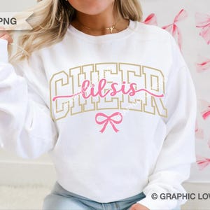 May include: White sweatshirt with the words "CHEER lil sis" in gold and pink lettering, with a pink bow graphic. The sweatshirt is a casual style, perfect for cheerleading or a sister. The background has a pattern of pink bows.