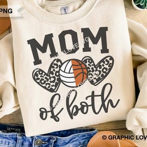 Pode incluir: Uma camisola de cor creme com o texto "MOM of both" em preto. O design inclui uma bola de voleibol com detalhes em laranja e branco, e dois corações com padrão de leopardo.