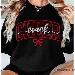 Peut inclure: Sweat-shirt noir avec le mot "CHEER" en lettres rouges et le mot "coach" en écriture blanche. Un nœud rouge est en dessous. Vêtement sur le thème du cheerleading ou de l'entraînement.