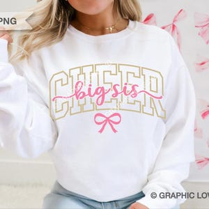 Puede incluir: Sudadera blanca con las palabras "CHEER big sis" en letras doradas y rosas, con un lazo rosa. Una prenda de estilo casual, perfecta para eventos de porristas o uso diario.