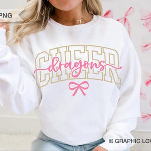 May include: White sweatshirt with the text "CHEER Dragons" in gold and pink lettering, with a pink bow graphic. The design is centered on the front of the shirt. The sweatshirt is a casual style.